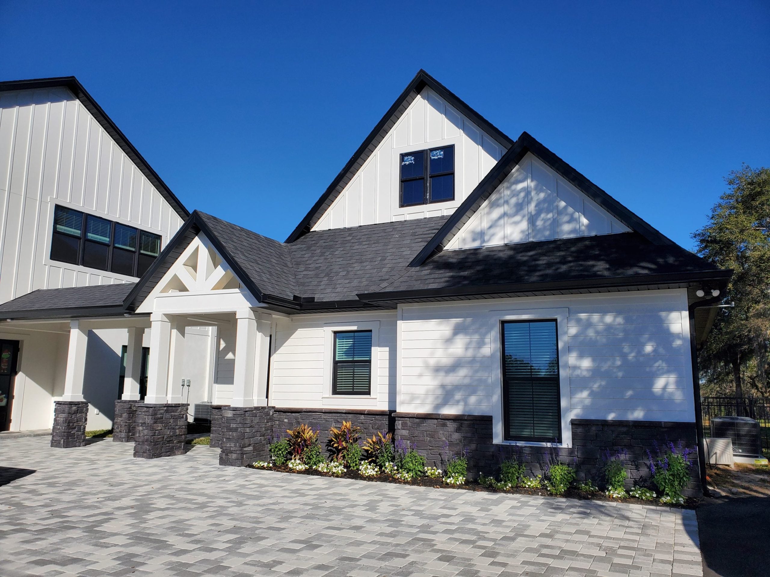 A modern white house with black trim, a gable roof, and a landscaped front yard with paved driveway under a clear blue sky.