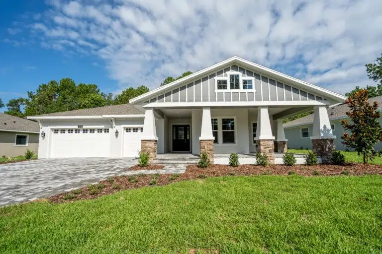 Single-story modern house with gray and white exterior, large front porch, two-car garage, and landscaped yard under a partly cloudy sky.