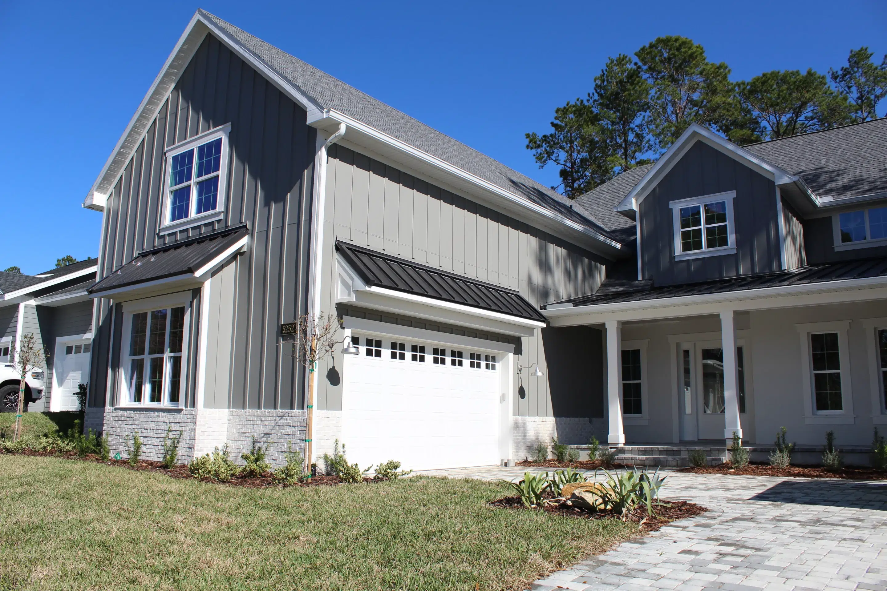 A modern gray two-story house with white trim, a white garage door, and a landscaped front yard under a clear blue sky.