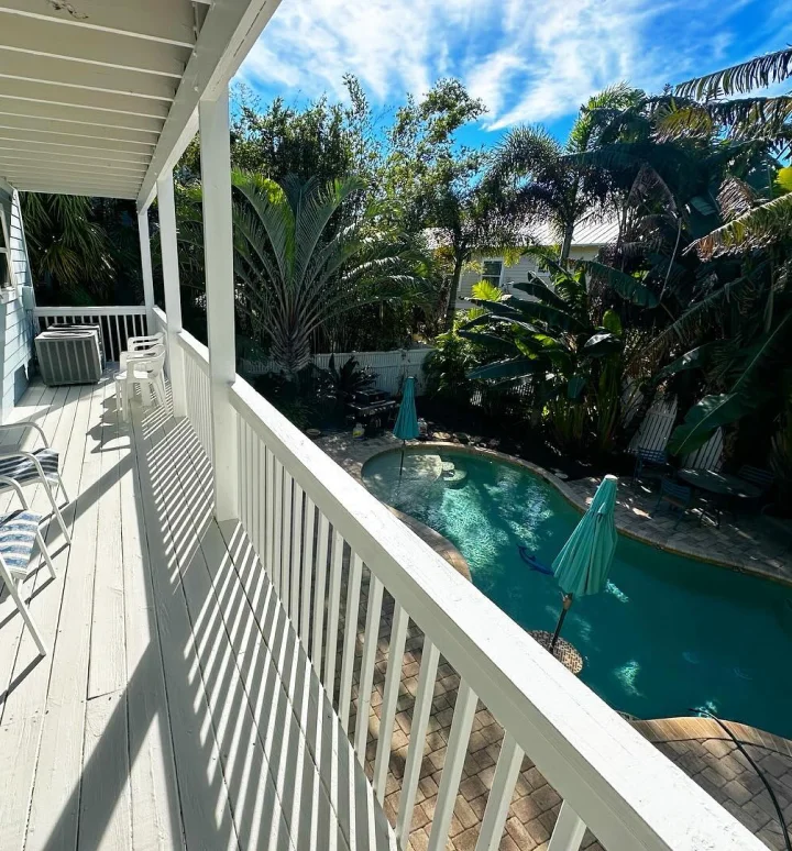 A sunlit balcony with white railing overlooks a backyard pool surrounded by tropical trees, lounge chairs, and closed umbrellas under a partly cloudy sky.